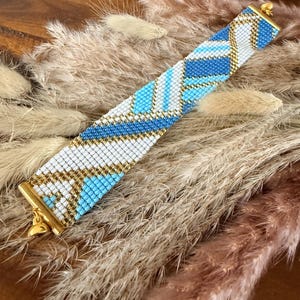 Puede incluir: Una pulsera de cuentas con un patrón geométrico en blanco, azul claro y dorado. La pulsera es rectangular y tiene un cierre dorado. Se muestra sobre un lecho de hierbas secas.