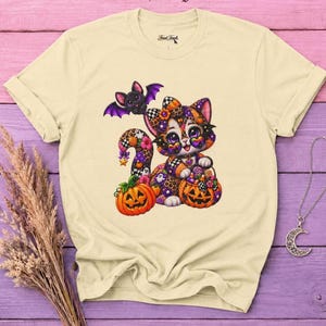 May include: A cream-coloured short-sleeve t-shirt featuring a graphic of a cat with large eyes, wearing a bow, and surrounded by two orange jack-o'-lanterns and a small purple bat. The cat has a patterned coat with orange, purple, and black colours.