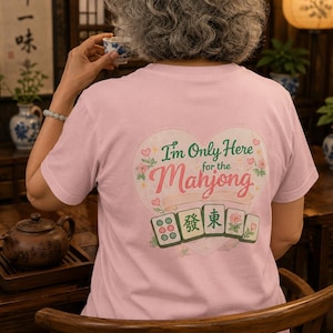 Puede incluir: Una camiseta de manga corta de color rosa claro con un gráfico grande en forma de corazón en la espalda. El diseño incluye el texto "I'm Only Here for the Mahjong" en letras verdes y rojas, rodeado de detalles florales y cuatro fichas de mahjong.