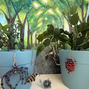 May include: A collection of wire and bead insect ornaments including a purple and black spider, a yellow and black bee, a red and black ladybug, and a small snail, all displayed on two light blue plastic pots with a forest painting background.