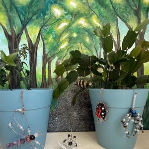 May include: Two light blue plastic plant pots containing green plants, decorated with wire and bead ornaments shaped like a dragonfly, a bee, a ladybug, a spider, and a snail. The ornaments feature various colorful beads and copper-colored wire.