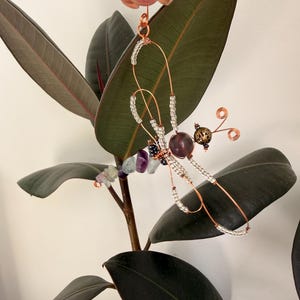 Può includere: Un ornamento a forma di libellula realizzato in filo di rame, con perline trasparenti lungo le ali, una perla centrale viola scuro, una perla metallica testurizzata per la testa e una coda composta da frammenti di pietre viola e verdi.