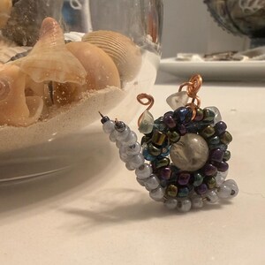 Peut inclure: Une petite figurine d'escargot fabriquée à partir de perles et de fil métallique. Le corps est composé de perles gris clair, tandis que la coquille présente une spirale de perles irisées violet foncé, vertes et bleues avec une perle transparente au centre. Deux antennes en fil de cuivre dépassent de la tête.
