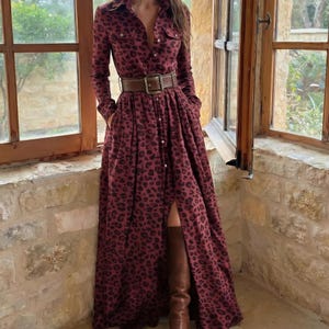 May include: A long, burgundy dress with a leopard print pattern. The dress features long sleeves, a button-down front, and a brown belt cinching the waist. Brown knee-high boots complete the look. The dress has two front pockets.