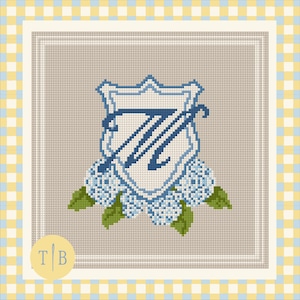 Puede incluir: Un patrón de punto de cruz sobre tela beige que presenta un escudo azul central con una letra W en cursiva. El escudo está enmarcado por racimos de flores azul claro y hojas verdes. Un logotipo circular con las letras T y B aparece en la esquina.