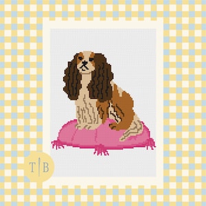 Könnte beinhalten: Ein Kreuzstichmuster mit einem Cavalier King Charles Spaniel mit braun-weißem Fell, der auf einem leuchtend pinken, getufteten Kissen sitzt. Der Hund hat lange, gewellte Ohren und einen konzentrierten Blick vor einem weißen Hintergrund.