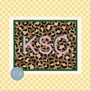 May include: A rectangular needlepoint canvas featuring a brown, black, and tan leopard print pattern. The center displays the letters KSC in light pink with a dark green outline. A dark green border frames the design, set against a yellow checkered background.