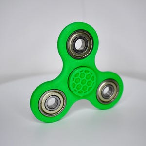 May include: A bright green fidget spinner with three weighted sections and silver bearings. The center section has a honeycomb pattern. The toy is designed to be spun between the fingers for stress relief or entertainment.