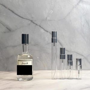 Peut inclure: Plusieurs flacons de parfum en verre transparent avec vaporisateurs argentés et bouchons noirs. Le plus grand flacon porte une étiquette avec le mot "fresh". Les flacons sont disposés en rangée, de tailles variées.