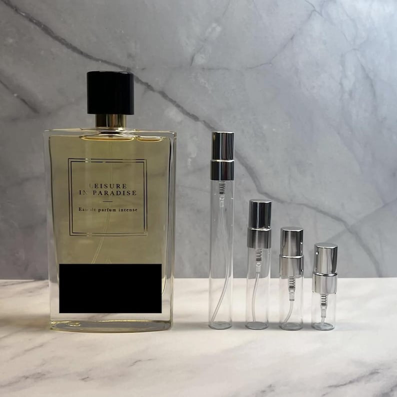 Puede incluir: Una botella de perfume rectangular con tapa negra y la inscripci&oacute;n "LEISURE IN PARADISE". Junto a la botella de perfume hay varios frascos de spray de vidrio transparente de varios tama&ntilde;os. La botella de perfume tiene una etiqueta rectangular negra.