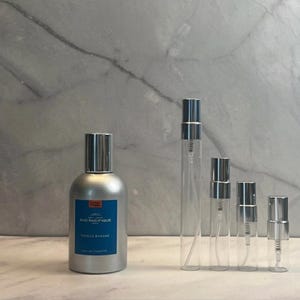 May include: A silver perfume bottle and four clear glass spray bottles with silver caps. The perfume bottle has a blue label with white text that reads "SUO PACIFIQUE VANILLE BANANE EAU DE TOILETTE."