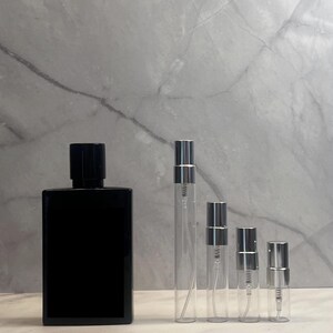 May include: A black rectangular perfume bottle with a black cap stands next to four clear glass spray bottles with silver caps. The bottles are arranged in a row against a marble background.
