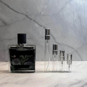 May include: A rectangular perfume bottle with a black cap and a black label featuring a floral design, next to four clear glass spray bottles with silver caps. The bottles are arranged on a marble surface.