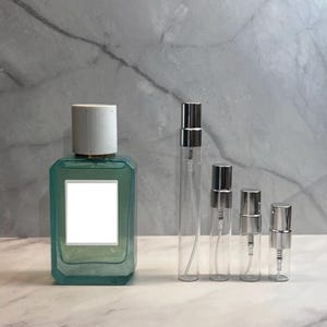 May include: A collection of perfume bottles and atomizers. A large, rectangular turquoise bottle with a white label is next to four clear glass atomizers with silver spray tops. The bottles are arranged on a marble surface.