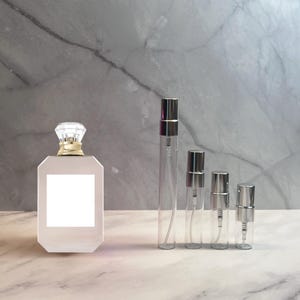 May include: A collection of perfume bottles and atomizers. The largest bottle is a rectangular shape with a clear, faceted stopper and a gold-colored neck. Four clear glass atomizers with silver-colored caps are lined up next to the larger bottle.