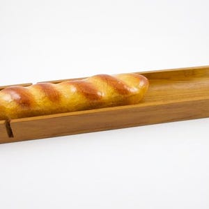 May include: A long, golden-brown baguette rests inside a rectangular, light brown wooden tray. The tray features a curved, trough-like interior and a small vertical notch cut into one of the side walls, showcasing a smooth, polished wood grain finish.