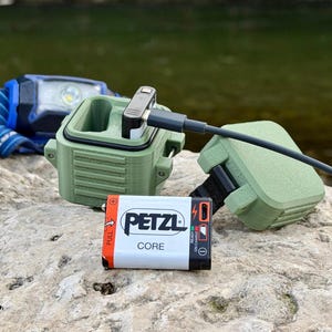 Op de afbeelding: Een oplaadbare Petzl Core batterij met een witte en oranje behuizing, geplaatst voor een olijfgroene plastic oplaadcase. Een zwarte USB-kabel is aangesloten op de batterij, die naast een blauwe en zwarte hoofdlamp ligt.