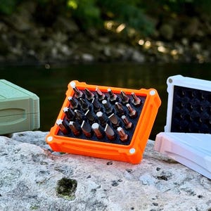 Op de afbeelding: Een set metalen schroevendraaierbits in een feloranje rechthoekige behuizing met een zwart schuimrubberen inzetstuk. Links staat een gesloten olijfgroene behuizing en rechts een witte behuizing met een zwarte binnenkant, beide getextureerd.