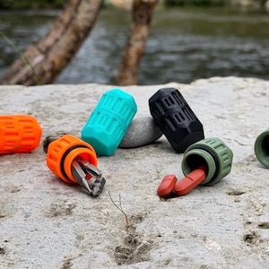Könnte beinhalten: Eine Sammlung kleiner, zylindrischer Schraubbehälter in Orange, Türkis, Schwarz und Olivgrün. Ein orangefarbener Behälter enthält Metall-Schraubendreher-Bits und ein olivgrüner Behälter enthält zwei rote Pillen. Die Behälter haben eine gerippte Oberfläche.