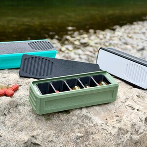 May include: Four rectangular pill organisers in teal, black, white, and olive green, each featuring a textured exterior and a ribbed top section. One olive green organiser is open, revealing five internal compartments containing various capsules.