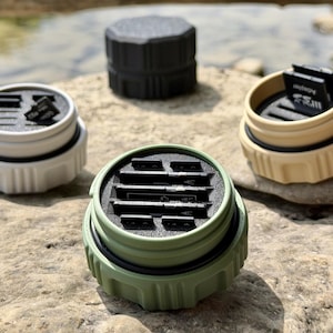 May include: Four circular, rugged storage cases for memory cards in black, white, tan, and olive green. Each case features a textured exterior, a black rubber O-ring seal, and an internal foam insert with slots designed to hold multiple SD and micro SD cards.