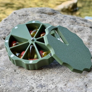 Può includere: Un portapillole rotondo verde oliva con otto scomparti a forma di spicchio contenenti varie capsule in gel rosse e gialle. Il contenitore presenta una superficie ruvida, un perno centrale e un coperchio rimovibile abbinato con maniglia.