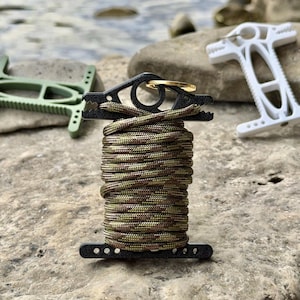 May include: A black plastic cord winder wrapped with camouflage paracord, featuring a gold-toned metal ring at the top. Two additional winders in olive green and white are positioned in the background, each with a distinct T-shaped design.