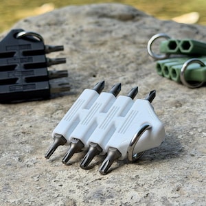 Könnte beinhalten: Drei Schraubendreher-Bit-Halter mit Metall-Schlüsselringen. Ein Halter ist schwarz, einer weiß und einer olivgrün. Jeder Halter fasst vier Metall-Schraubendreher-Bits in einer Reihe und verfügt über einen strukturierten Griff und ein kompaktes Design.