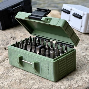 Può includere: Una custodia rettangolare verde oliva con coperchio incernierato e chiusura nera, contenente una collezione di vari inserti per cacciavite in metallo. La custodia presenta un esterno testurizzato e una piccola maniglia in metallo.