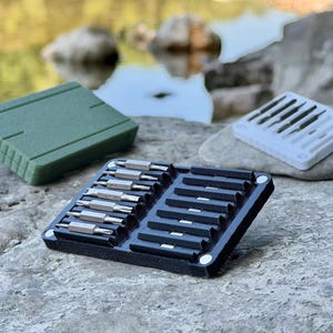 Op de afbeelding: Een zwarte rechthoekige organizer-tray met zes dubbelzijdige metalen schroevendraaierbits, inclusief een bijpassende olijfgroene gesloten etui en een witte tray met extra bits geplaatst op een gestructureerd stenen oppervlak.