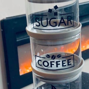 Könnte beinhalten: Ein Set aus drei transparenten Glasbehältern mit grauen Deckeln, beschriftet mit "ZUCKER", "KAFFEE" und "TEE" in schwarzer Schrift. Die Behälter sind gestapelt, mit einem Kamin im Hintergrund.