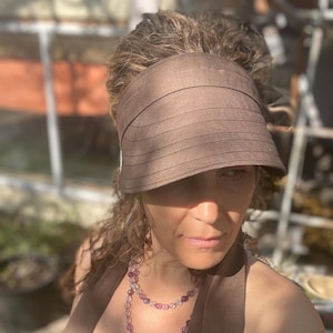 May include: A woman wearing a brown sun visor and a layered necklace of purple and pink beads. She is wearing a brown tank top and a brown scarf.
