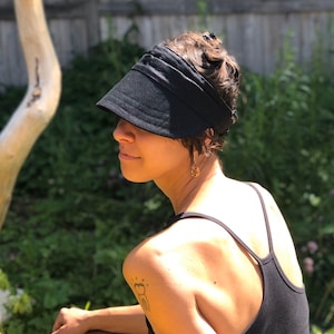 May include: A black visor with a wide brim, worn on the head. The visor is made of a soft, dark fabric. The person is wearing a black vest top. The background is a natural outdoor setting with greenery and a wooden fence.