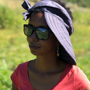 Può includere: Una donna con un copricapo blu polveroso legato con un fiocco e occhiali da sole neri. Indossa anche una maglietta con scollo a V color corallo e orecchini. Lo sfondo è un campo verde sfocato.