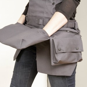 Può includere: Un grembiule grigio con più tasche e un ampio lembo che può essere ripiegato per creare una tasca. Il grembiule ha una cintura e una chiusura a zip.