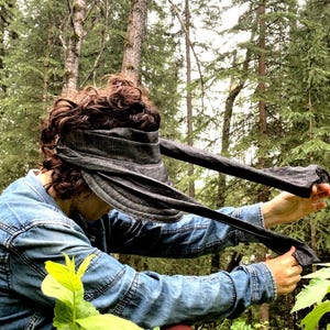 May include: A person wearing a dark gray denim bandana as a headband and a denim jacket. The bandana is tied around their head and the ends are flowing in the wind. The person is standing in a forest.
