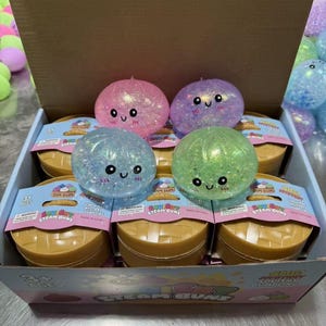 Puede incluir: Una caja de cartón llena de juguetes Dim Sum Steam Buns. Los juguetes son redondos, coloridos y tienen caras bonitas. Los bollos están en un recipiente marrón con una etiqueta azul y blanca. La parte superior de la caja tiene varios juguetes redondos y brillantes.