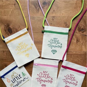 May include: Five small, white canvas crossbody bags with colourful zips and cords. Each bag features text and a graphic: "MY Little Treasures" with a diamond, and "LITTLE explorer" with a mountain and tree. Zips are gold, green, pink, and red.
