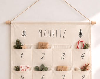 Könnte beinhalten: Ein cremefarbener Wand-Adventskalender aus Stoff mit vierundzwanzig Taschen, die jeweils mit schwarzer Tinte nummeriert sind. Der Name MAURITZ ist oben zwischen zwei Baum-Symbolen aufgedruckt. Der Kalender hängt an einem Holzstab.