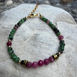 Könnte beinhalten: Ein Perlenarmband mit facettierten grünen und magentafarbenen Edelsteinperlen, durchsetzt mit kleinen goldfarbenen Metallzwischenperlen und zylindrischen goldfarbenen Akzenten. Das Armband hat einen goldfarbenen Karabinerverschluss und eine verstellbare Kette.