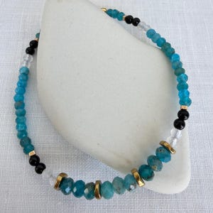 Könnte beinhalten: Ein Armband mit türkisfarbenen, schwarzen und klaren Perlen, unterbrochen von goldfarbenen Elementen. Das Armband wird auf einem weißen Stein präsentiert, der seine Farben und sein Design hervorhebt. Dieses Schmuckstück ist für den täglichen Gebrauch geeignet, ca. 18 cm lang.