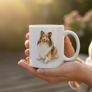 Peut inclure: Mug en céramique blanche avec une illustration à l'aquarelle d'un chien Sheltie. Le chien a des poils bruns, blancs et bronzés, avec une expression joyeuse. La tasse a une anse incurvée et est tenue dans les mains d'une personne.