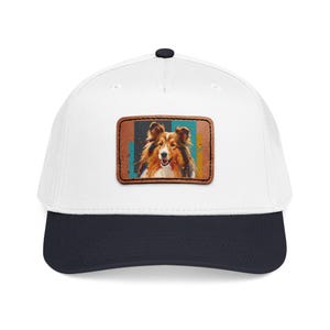 Può includere: Un cappellino da baseball bianco con visiera blu navy e bottone nero. Il cappellino presenta una toppa rettangolare con un dipinto colorato di un cane Sheltie. Il cane ha pelo marrone e bianco.