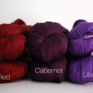 May include: A collection of colorful yarn skeins in shades of red, purple, and lilac. The image features three distinct colors: Red, Cabernet, and Lilac. Each skein is labeled with its respective color name, offering a clear visual guide for crafters.