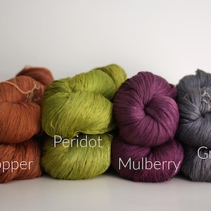 May include: A collection of yarn skeins in various colors: copper, peridot, mulberry, and gray. The yarn is arranged in a stack, with the color names labeled on the image. The yarn appears soft and fluffy, ideal for knitting or crocheting projects.