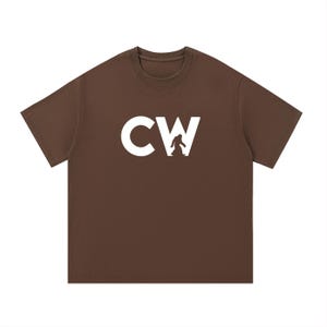 May include: A brown t-shirt with the white letters "CW" and a white silhouette of a Bigfoot creature. The shirt has a crew neck and short sleeves. The image is on a white background.