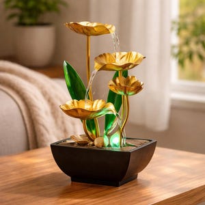 May include: An indoor water feature with a tiered design. The fountain has golden lotus-shaped bowls with water flowing into a black square base. Green leaves and a small green light enhance the look. The fountain is placed on a wooden table.
