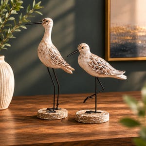 May include: Two decorative bird figurines. The birds have a cream-colored body with brown detailing on the wings. They stand on a round, textured base with black metal legs. The birds have long black beaks and are positioned on a wooden surface.