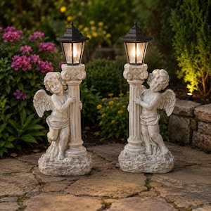 Puede incluir: Dos lámparas de jardín exteriores con faroles negros y estatuas de querubines. Las lámparas están sostenidas por columnas ornamentadas y se encuentran en un patio de piedra. Los querubines tienen alas.
