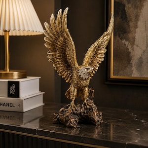 May include: A gold-colored eagle statue with outstretched wings, perched on a textured, brown base. The eagle's feathers are intricately detailed, and its head features silver and white accents. The statue is a decorative item.